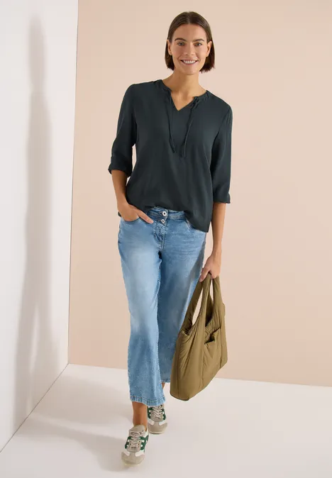 Vrouw voor lichte achtergrond met donker topje, blauwe jeans, sneakers en een groene schoudertas.