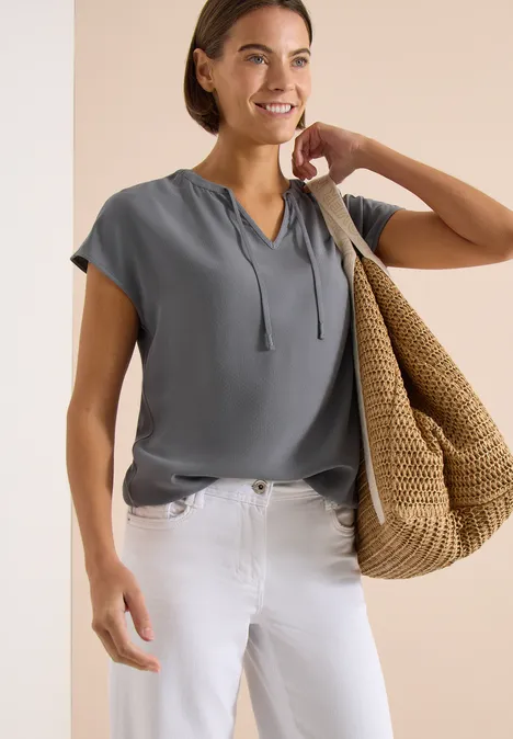 Vrouw in grijze korte mouw top, witte jeans, draagt grote stroentas op haar schouder voor een beige achtergrond.