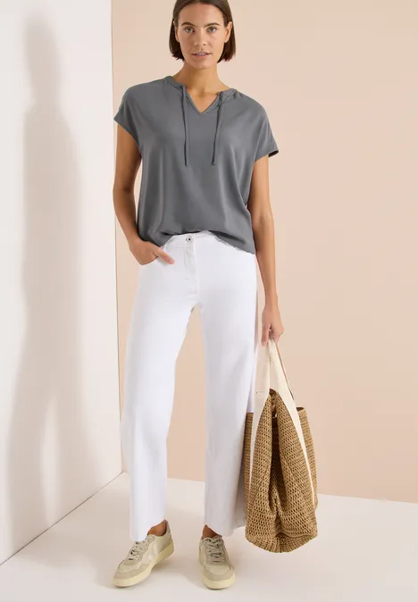 Vrouw in grijs shirt, witte broek en strooien tas voor een beige muur.