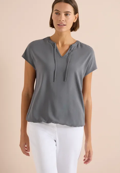 Vrouw in grijs kort mouw capuchonshirt met V-hals en witte broek.