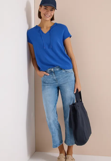 Persoon met blauwe blouse, spijkerbroek en pet, draagt blauwe tas en leunt tegen muur.