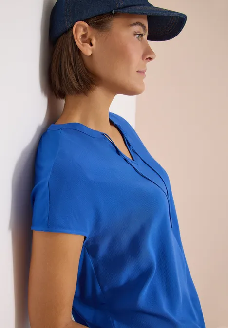 Vrouw met blauw kort mouwshirt en donkerblauw petje leunt tegen een lichte muur.
