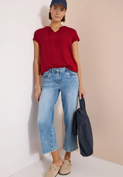 Vrouw in rode blouse, wijde lichtblauwe jeans en beige schoenen, met een donkerblauwe tas.