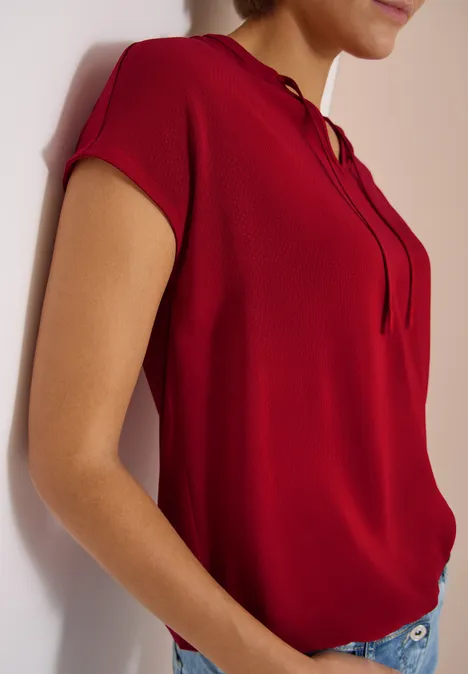 Een vrouw draagt een rood shirt met korte mouwen en jeans terwijl ze tegen een muur leunt.