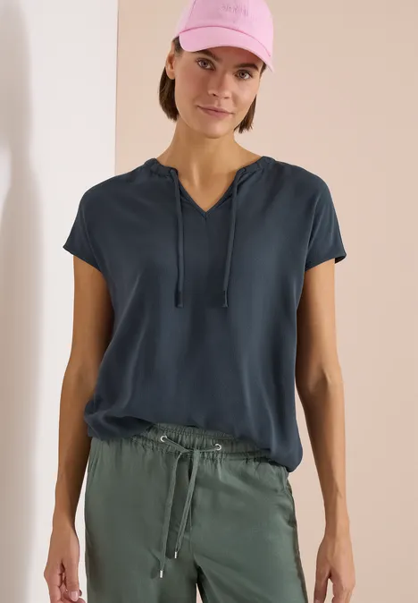 Een persoon draagt een donkerblauw shirt met korte mouwen en koordjes, gecombineerd met een olijfgroene broek.
