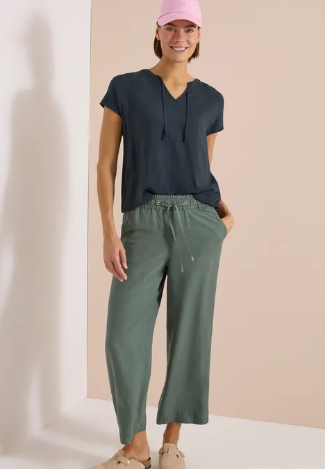 Persoon met zwart V-hals top en loszittende olijfgroene broek voor een lichte achtergrond.