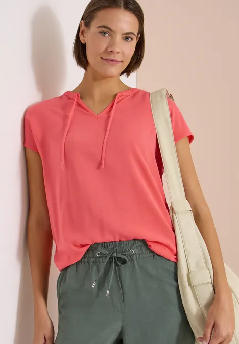 Vrouw staat tegen lichte muur, draagt roze korte mouw hoodie, groene broek en een beige katoenen tas.