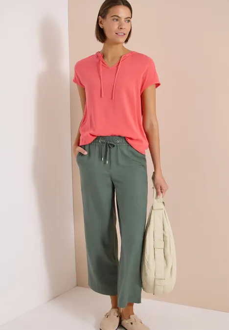 Vrouw in roze T-shirt met V-hals, groene wijde broek tot op de enkel en draagt een witte tas.