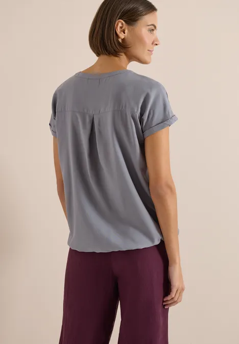 Blouse met korte mouwen en gespleten hals graphite light grey Vrouw met rug naar camera, draagt een grijs T-shirt met korte mouwen en paarse broek tegen een lichte achtergrond.