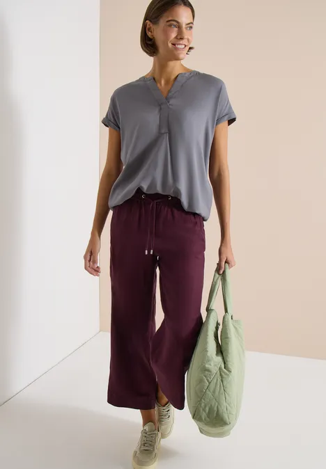 Blouse met korte mouwen en gespleten hals graphite light grey Een vrouw draagt wijde paarse broek, een grijs T-shirt met korte mouwen, beige sneakers en een lichtgroene tas.