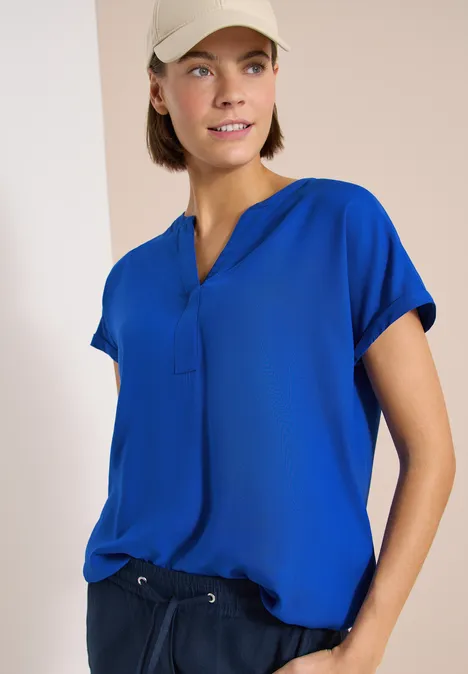 Blouse met korte mouwen en gespleten hals surf blue Een persoon draagt een blauwe blouse met korte mouwen en een donkere broek tegen een neutrale achtergrond.