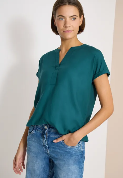 Blouse met korte mouwen en gespleten hals jungle green Vrouw draagt donkergroen kort mouw blouse met knoopsluiting en blauwe spijkerbroek.