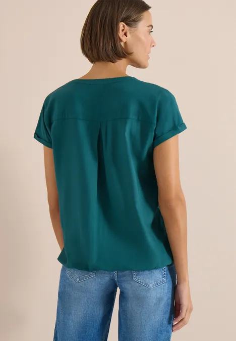 Blouse met korte mouwen en gespleten hals jungle green Een vrouw van achteren in een groen t-shirt en een blauwe spijkerbroek.