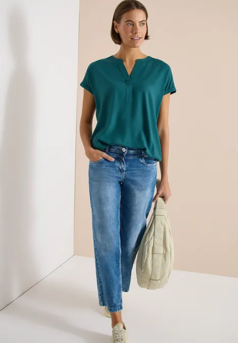 Blouse met korte mouwen en gespleten hals jungle green Vrouw draagt groen V-hals shirt, blauwe jeans en grijze sneakers, houdt beige gewatteerde tas vast.