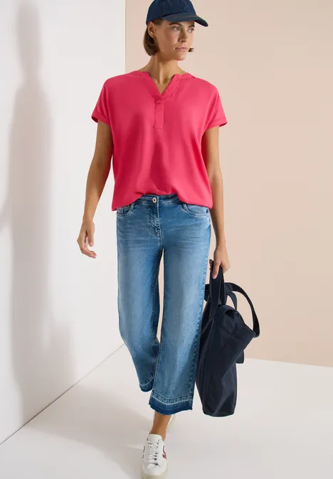 Blouse met korte mouwen en gespleten hals geranium red Persoon in roze shirt, blauwe jeans en witte sneakers, met een donkerblauwe tas.