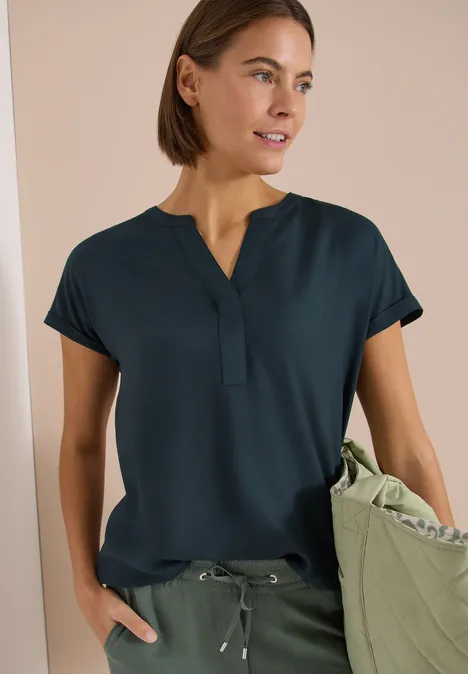 Kurzarm Bluse mit Split Neck deep petrol blue Frau trägt dunkel-türkises Kurzarmshirt mit V-Ausschnitt, olivgrüne Hose mit Kordelzug und passende Tasche.
