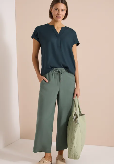 Kurzarm Bluse mit Split Neck deep petrol blue Frau mit dunkelblauem Shirt, grünen, weiten 7/8-Hosen und passender, gesteppter Umhängetasche vor beigem Hintergrund.