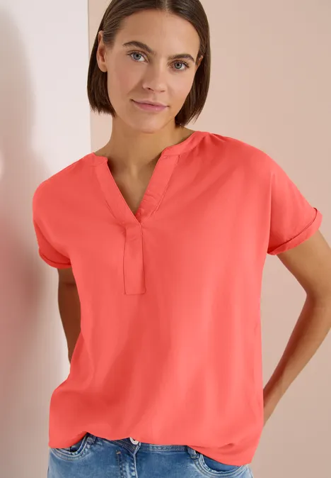 Kurzarm Bluse mit Split Neck shiny coral Eine Frau trägt eine korallenrote, kurzärmelige Bluse mit V-Ausschnitt vor hell beigem Hintergrund.