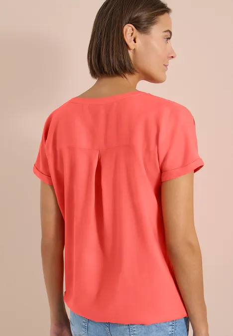 Kurzarm Bluse mit Split Neck shiny coral Frau von hinten in korallfarbener Bluse mit Kellerfalte vor neutralem Hintergrund