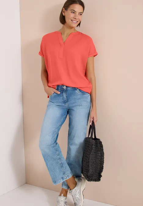 Kurzarm Bluse mit Split Neck shiny coral Model trägt korallrotes Shirt, blaue Jeans, weiße Sneaker und hält eine schwarze Flecht-Tasche.