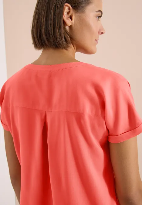 Kurzarm Bluse mit Split Neck shiny coral Rückansicht einer Person in einem korallenroten Hemd vor hellem Hintergrund.