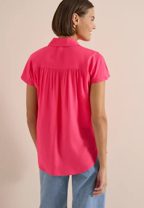 Vrouw van achteren in een felroze blouse met korte mouwen en kraag, gecombineerd met een spijkerbroek.