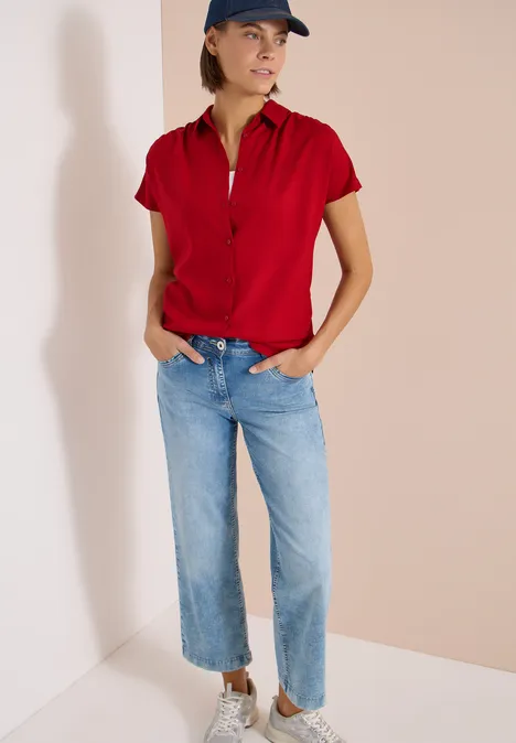 Frau in rotem Polohemd und blauen Jeans, Hände in den Taschen, steht entspannt in einer Ecke vor hellem Hintergrund.