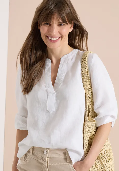 3/4-Arm Tunika aus reinem Leinen White Frau in weißer Leinenbluse, beiger Hose und Strohtasche vor neutralem Hintergrund.