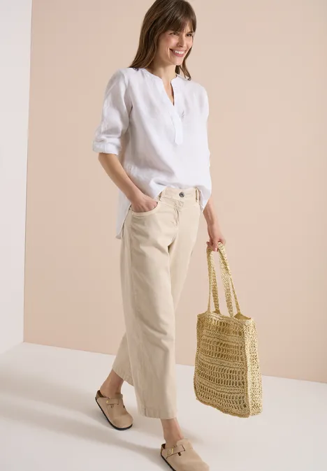 3/4-Arm Tunika aus reinem Leinen White Frau in weißer Leinenbluse, beigen Cropped-Hosen, geflochtenen Espadrilles und Strohtasche vor hellem Hintergrund.