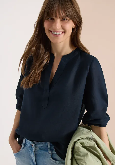 3/4-Arm Tunika aus reinem Leinen universal blue Frau in dunkelblauer Bluse und Jeans, hält eine grüne Jacke in der Hand.