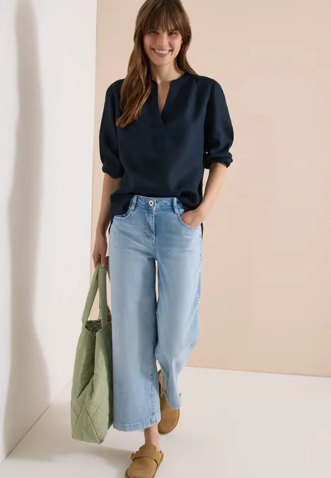 3/4-Arm Tunika aus reinem Leinen universal blue Frau in lässiger Bluse und weiten Jeans lehnt entspannt an Wand, trägt grüne Stofftasche.