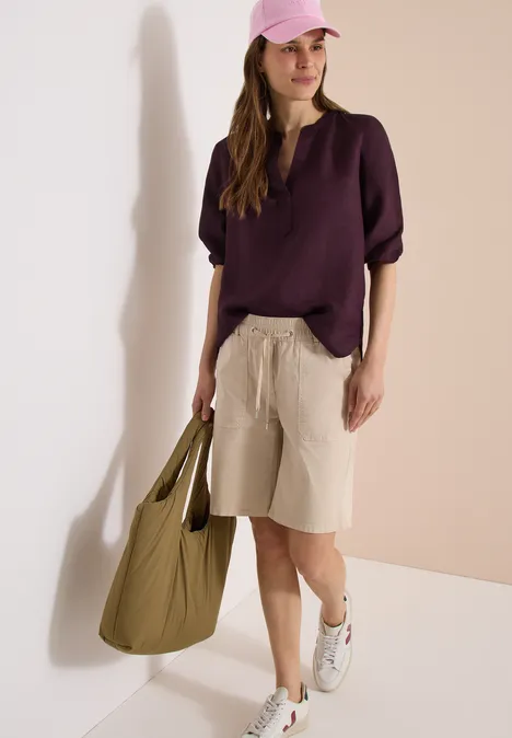 Femme en chemise violette, bermuda beige, baskets blanches et grand sac vert sur fond neutre.