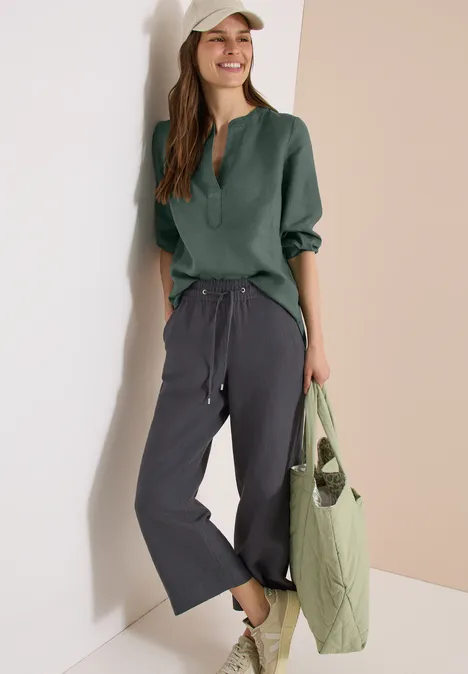 3/4-Arm Tunika aus reinem Leinen just khaki Person lehnt an Wand, trägt grünes Langarmhemd, weite graue Hose und gesteppte grüne Tasche.