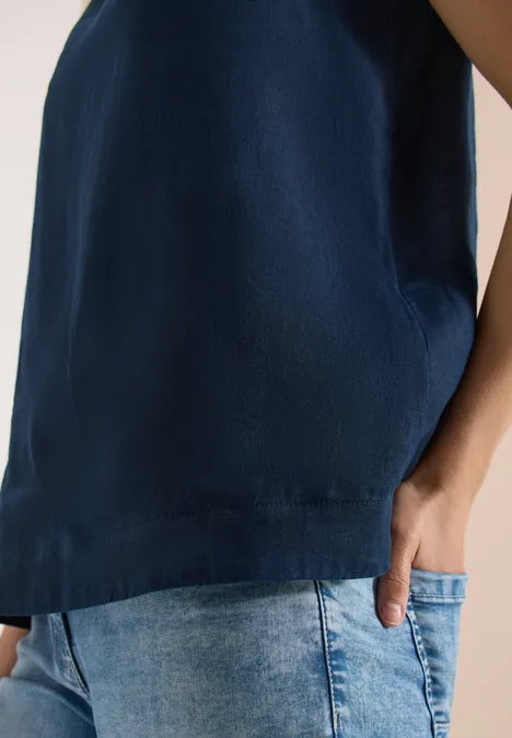 Eine Person trägt ein ärmelloses, dunkelblaues Oberteil und helle Jeans, Hand in der Tasche.