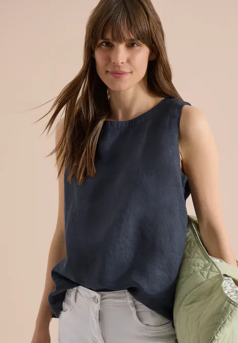 Frau trägt ärmelloses dunkelblaues Top, helle Hose und hält eine grüne Jacke in der Hand.