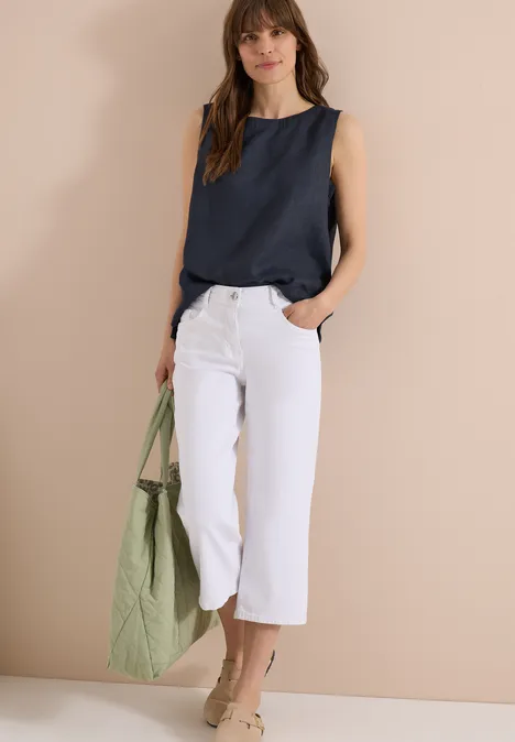 Frau trägt dunkelblaues Top, weiße Culotte und hält eine grüne Umhängetasche.