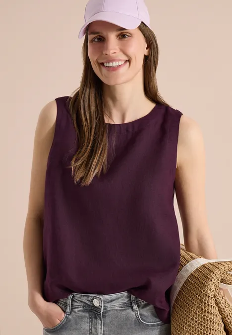 Modell trägt ärmellose, dunkelrote Bluse zu Jeans, hält Strohtasche