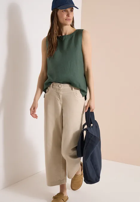 Frau mit grünem Top und beigen Weitbein-Hosen, trägt Mütze und dunkelblaue Tasche.