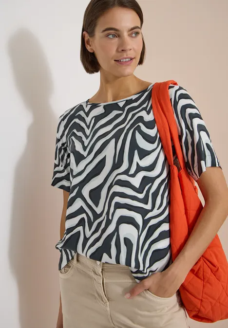Person trägt schwarz-weißes Zebra-Shirt, beige Hose und orangefarbene Tasche vor hellem Hintergrund.