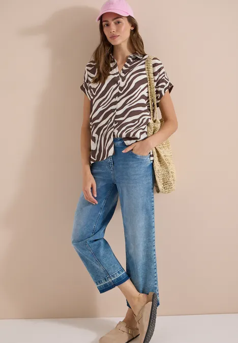 Kurzarm Hemdbluse mit Zebra-Print brown cacao Frau trägt Zebra-Print-Bluse, weite Jeans, Strohtasche, Espadrilles und Cap vor beiger Wand.
