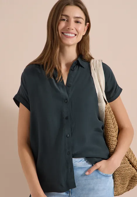 Kurzarm Hemdbluse in Unifarbe palm breeze green Frau trägt dunkelgrüne, kurzärmlige Bluse und hält Korbtasche, Hand in Hosentasche, vor hellem Hintergrund.