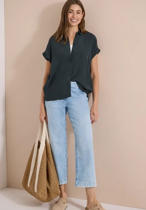Kurzarm Hemdbluse in Unifarbe palm breeze green Frau vor beigem Hintergrund trägt schwarze Bluse, hellblaue Culotte-Jeans und großen Strohtasche.