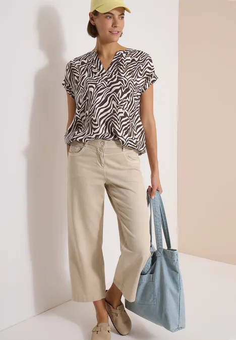 Frau in beiger Culottehose, Zebra-Musterbluse, lässigen Schuhen und blauer Stofftasche.