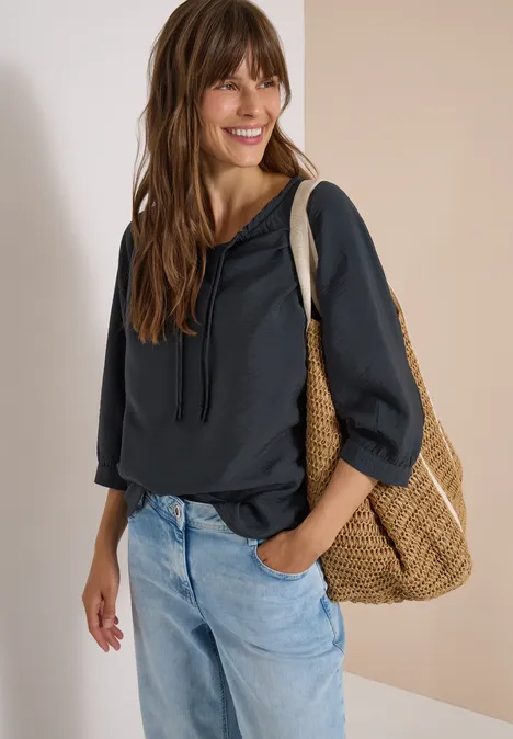 Chemisier Carmen à manches courtes et rubans palm breeze green Frau in dunkler Kapuzenbluse und hellen Jeans mit Korbtasche auf dem Arm, lächelt vor hellem Hintergrund.