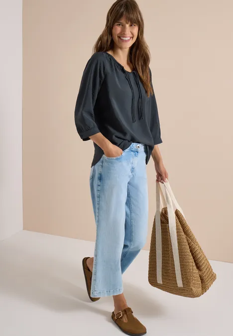 Chemisier Carmen à manches courtes et rubans palm breeze green Frau in dunkelblauer Bluse, hellen Jeans und Loafers trägt geflochtene Tasche und lächelt vor beigem Hintergrund.