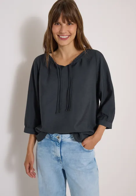 Chemisier Carmen à manches courtes et rubans palm breeze green Lächelnde Frau trägt dunkle Bluse mit Kordeln und helle Jeans, steht entspannt vor heller Wand.