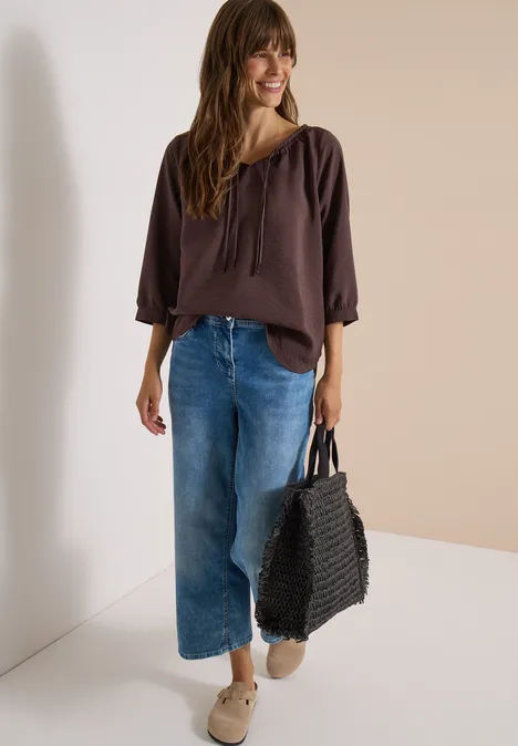 Chemisier Carmen à manches courtes et rubans brown cacao Frau in brauner Bluse, weiten Jeans und Sneakers trägt schwarze geflochtene Tasche und lächelt.