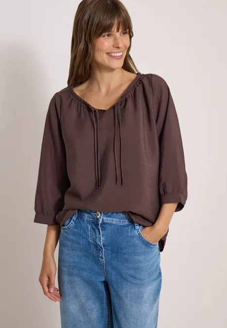 Chemisier Carmen à manches courtes et rubans brown cacao Lächelnde Frau in brauner Bluse und blauen Jeans, eine Hand in der Hosentasche.