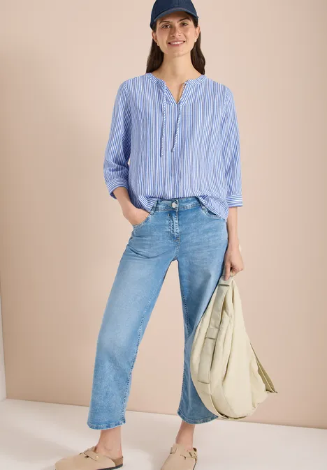 Vrouw in lichtblauw-wit gestreept overhemd, los in rechte jeans gestopt, met grote beige stoffen tas in de hand.