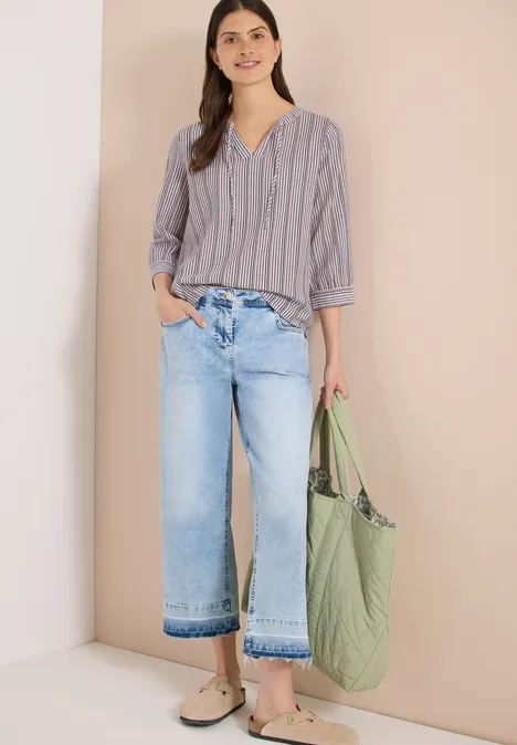 Vrouw in gestreept overhemd, wijde cropped jeans en groene tas voor een neutrale achtergrond.